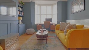 Ein Zimmer mit einem gelben Sofa, einem runden Couchtisch, einem Schreibtisch mit Stuhl und einem Fenster mit Jalousien.