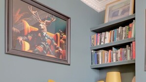 Ein Zimmer mit einem gelben Sessel, einem Bücherregal mit Büchern, einem gerahmten Bild an der Wand und einer Lampe auf einem Beistelltisch.