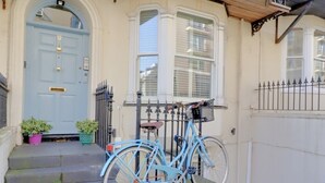 Ein blaues Fahrrad steht vor einem Gebäude mit einer weißen Tür, einem Balkon mit schmiedeeisernem Geländer und einem kleinen Gartenbereich mit Blumentöpfen.