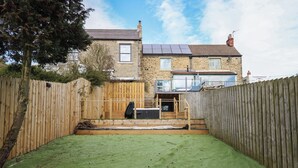 A backyard with artificial grass, a wooden deck, a hot tub, and a stone house with solar panels. - 2 Low Road (Bishop Auckland)