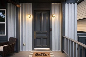 A dark grey door with two wall-mounted lights, a wicker chair, and a 'hello' mat on the porch. - Luxury Stay • Near Heights, Galleria & Downtown (Houston)