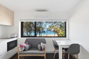 A modern kitchen with a view of a beach, a grey sofa, and a dining table with chairs. - Modern Beachside Studio (Blackmans Bay)
