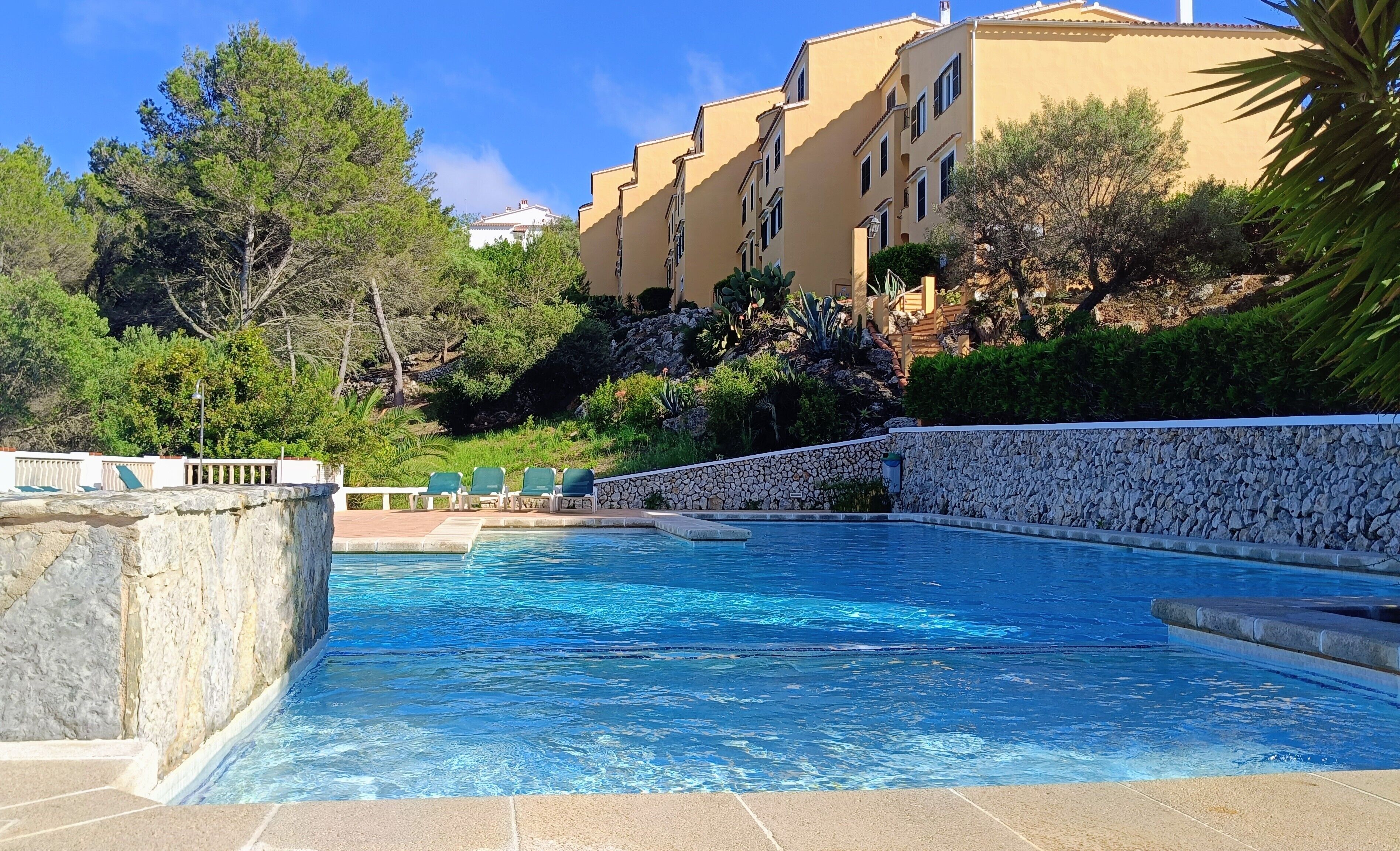 A swimming pool with a stone wall, green lounge chairs, and a building in the background.