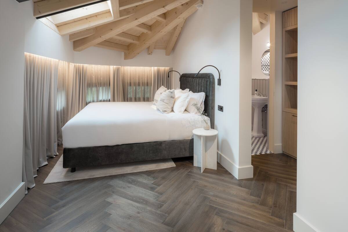 Une chambre avec un grand lit, des poutres apparentes, une lucarne, une table de chevet et une vue sur la salle de bain.