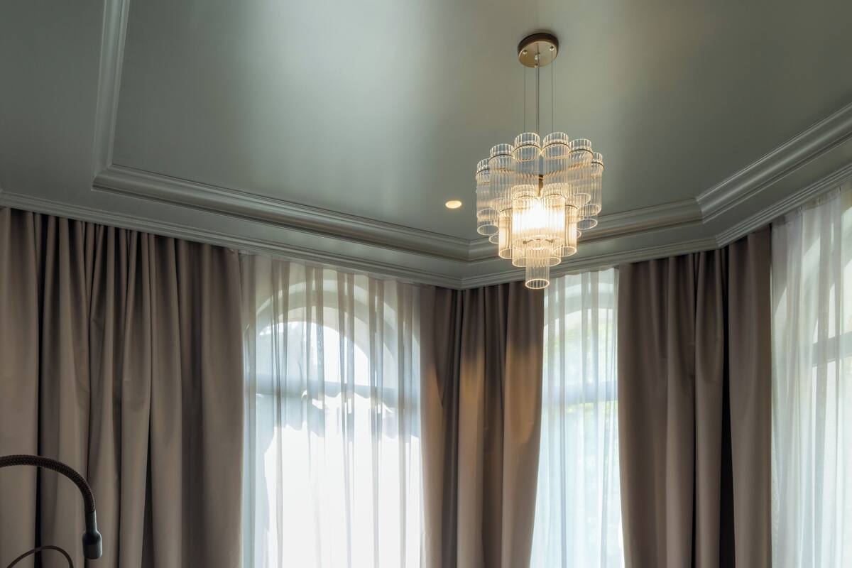 Un lit bien fait, avec du linge de lit blanc et une tête de lit recouverte d’un tissu vert, un lustre et de fines rideaux aux grandes fenêtres.