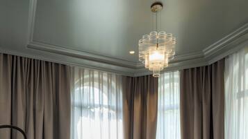 Cama bem arrumada com lençóis brancos e cabeceira estofada verde, um candelabro e cortinas leves em janelas grandes.