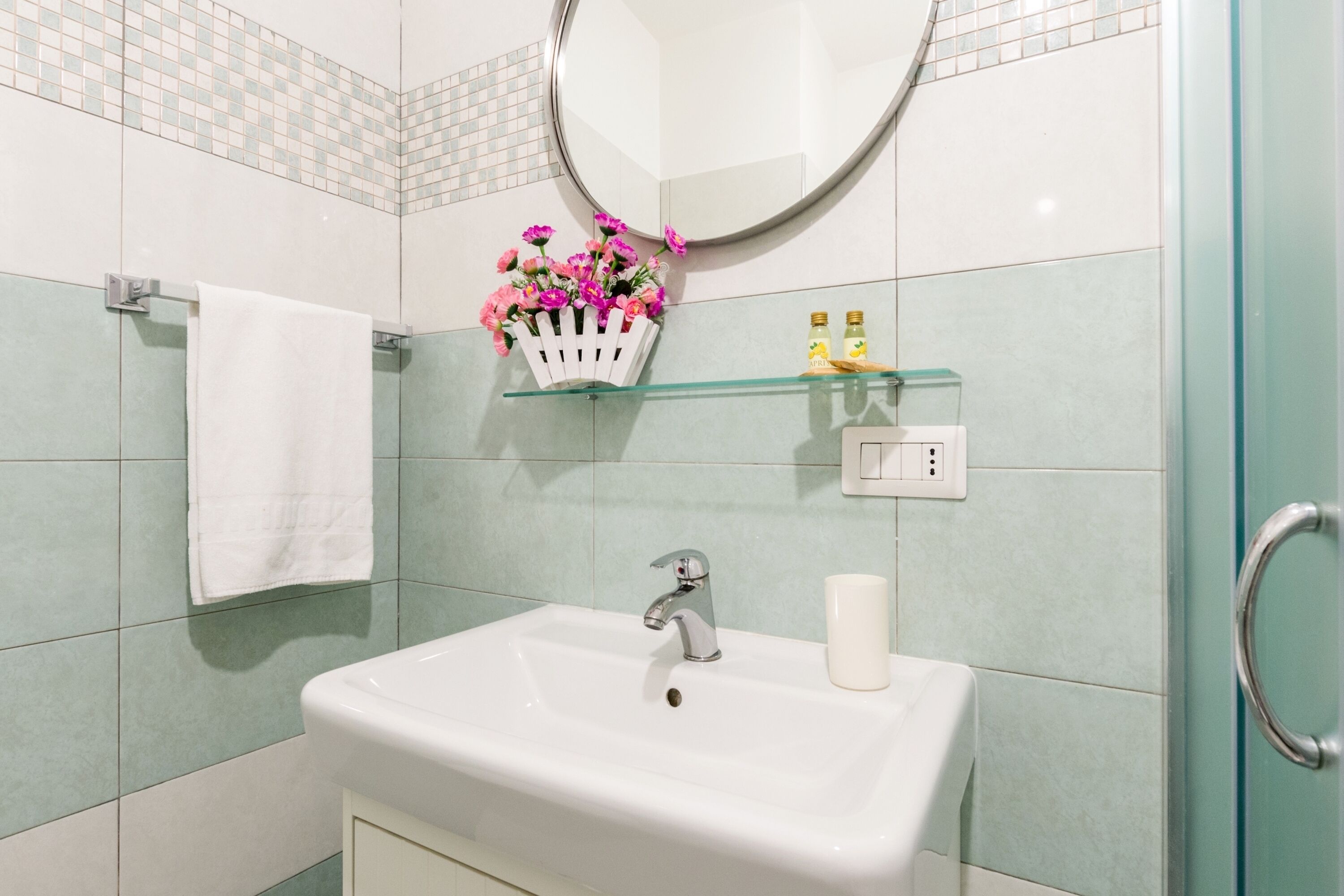 A bathroom with a white sink, a mirror, a white towel, a glass shelf with flowers, and a glass shower door.