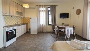 Uma cozinha compacta com fogão, geladeira e painel de fundo xadrez. Há uma área de jantar com mesa e cadeiras, e uma TV fixada na parede.
