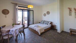 Quarto com cama, mesa e cadeiras, porta de varanda e pequena mesa com planta.