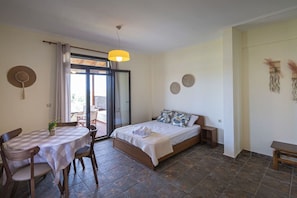 A bedroom with a bed, a table with chairs, a balcony door, and a small table with a plant.