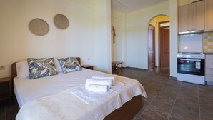 Quarto de hotel com cama, almofadas, uma pequena mesa, decoração na parede e uma área de cozinha com forno.