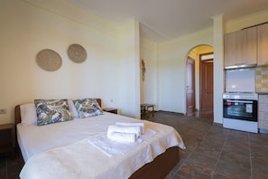 A hotel room with a bed, pillows, a small table, a wall decoration, and a kitchen area with an oven.