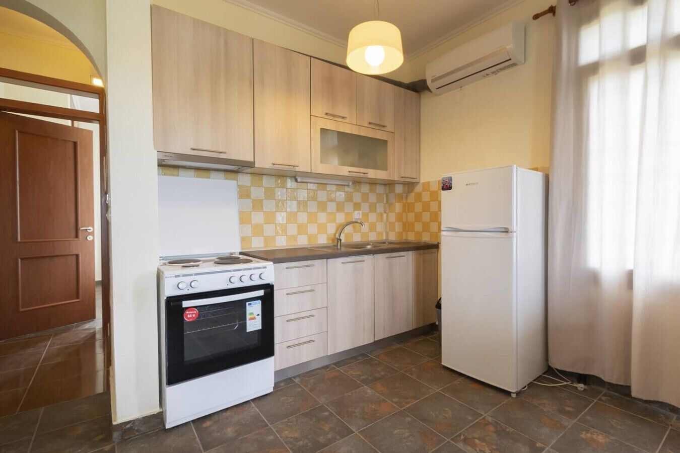 A compact kitchen with a stove, oven, sink, and refrigerator, light wooden cabinets, and a checkered backsplash.