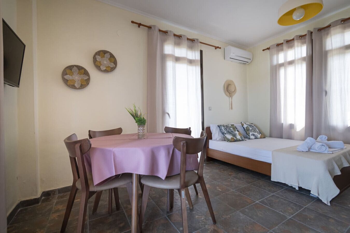 A room with a dining table, chairs, a bed, and a small table with a plant.