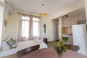 A hotel room with a bed, a desk, a chair, a dining table, a kitchen area, and a hanging plant. - Tres Flores Kalmia by RentalsPro - Agios Mamas Halkidiki (Agios Mamas)