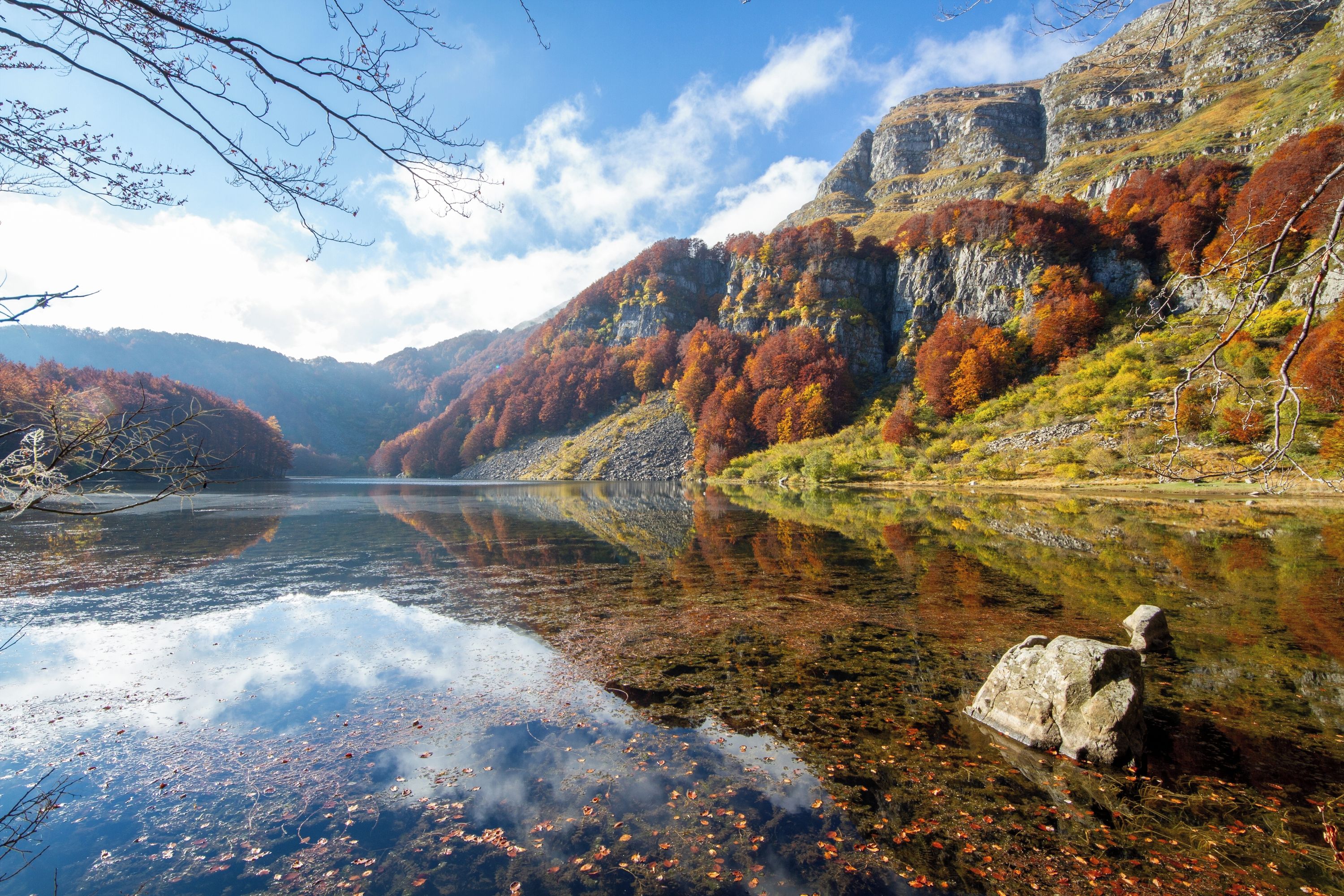 A serene lake with clear reflections, surrounded by autumn-colored trees and rocky cliffs.