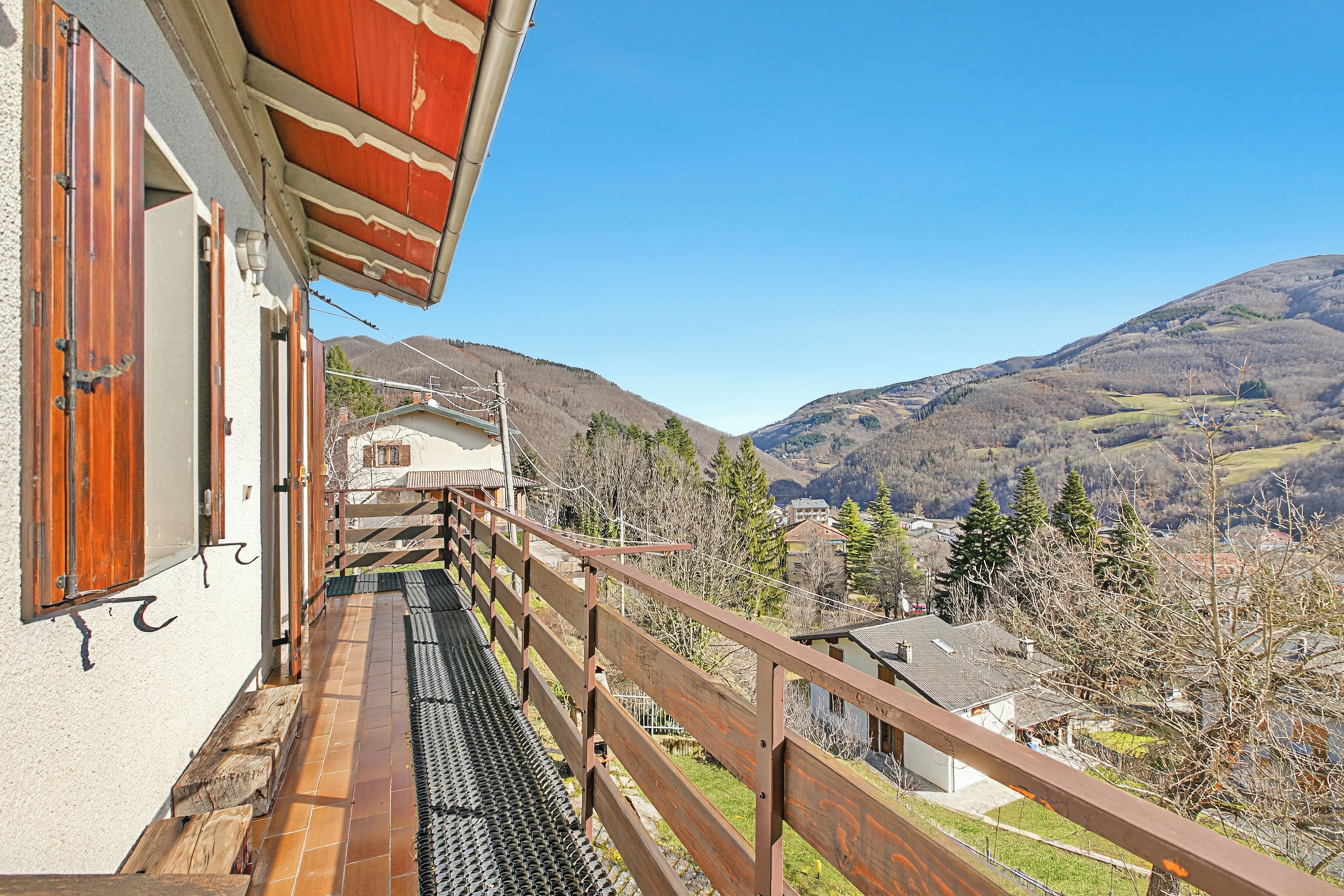 A balcony with a view of a mountainous landscape, a wooden railing, and a building with a sloped roof.
