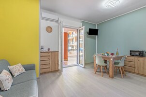 Una habitación con sofá, una mesa de comedor con sillas, un televisor montado en la pared y vistas a un balcón con barandillas.