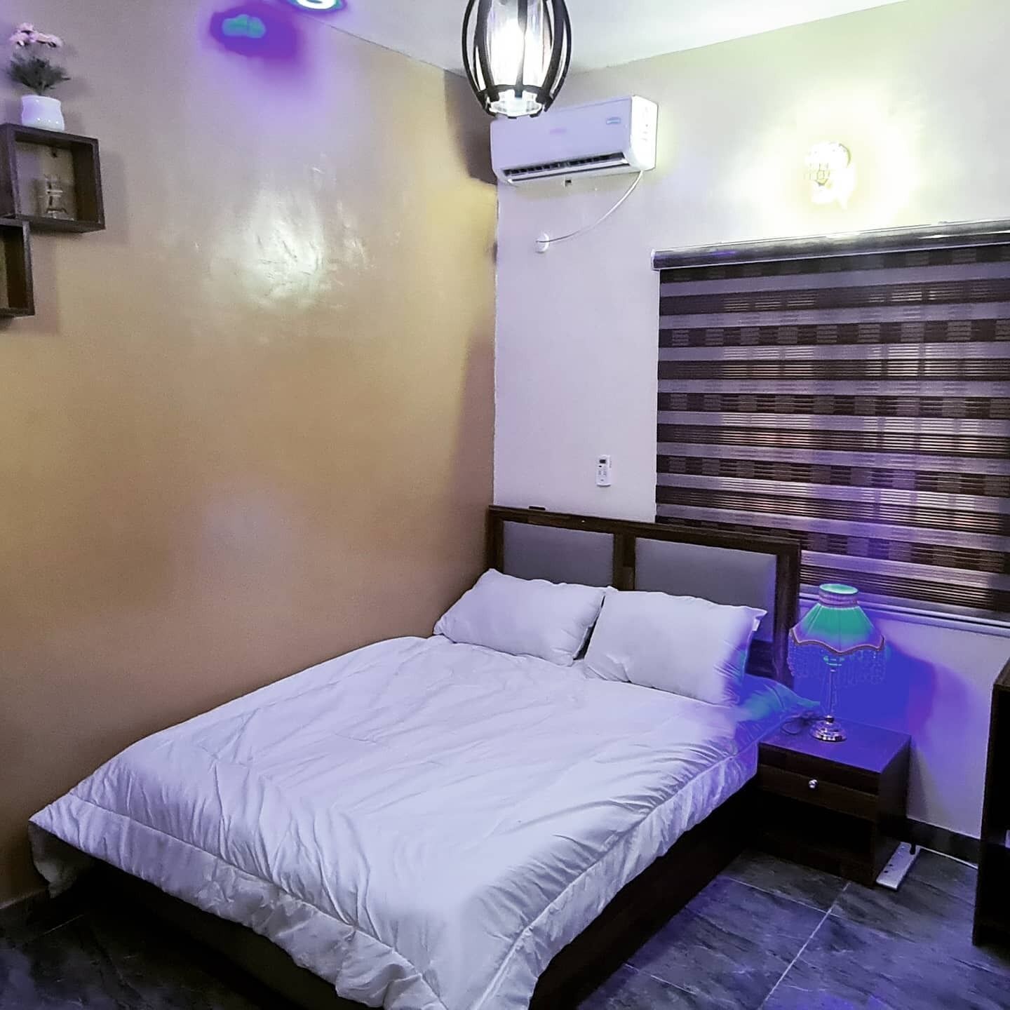 A bedroom with a bed, a nightstand, a lamp, an air conditioner, and a wall-mounted shelf with decorative items.