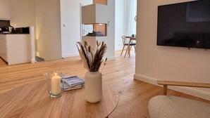 Een moderne woonkamer met een houten vloer, een glazen tafel met een kaars, een vaas met gedroogde planten, een bank en een aan de muur bevestigde televisie.