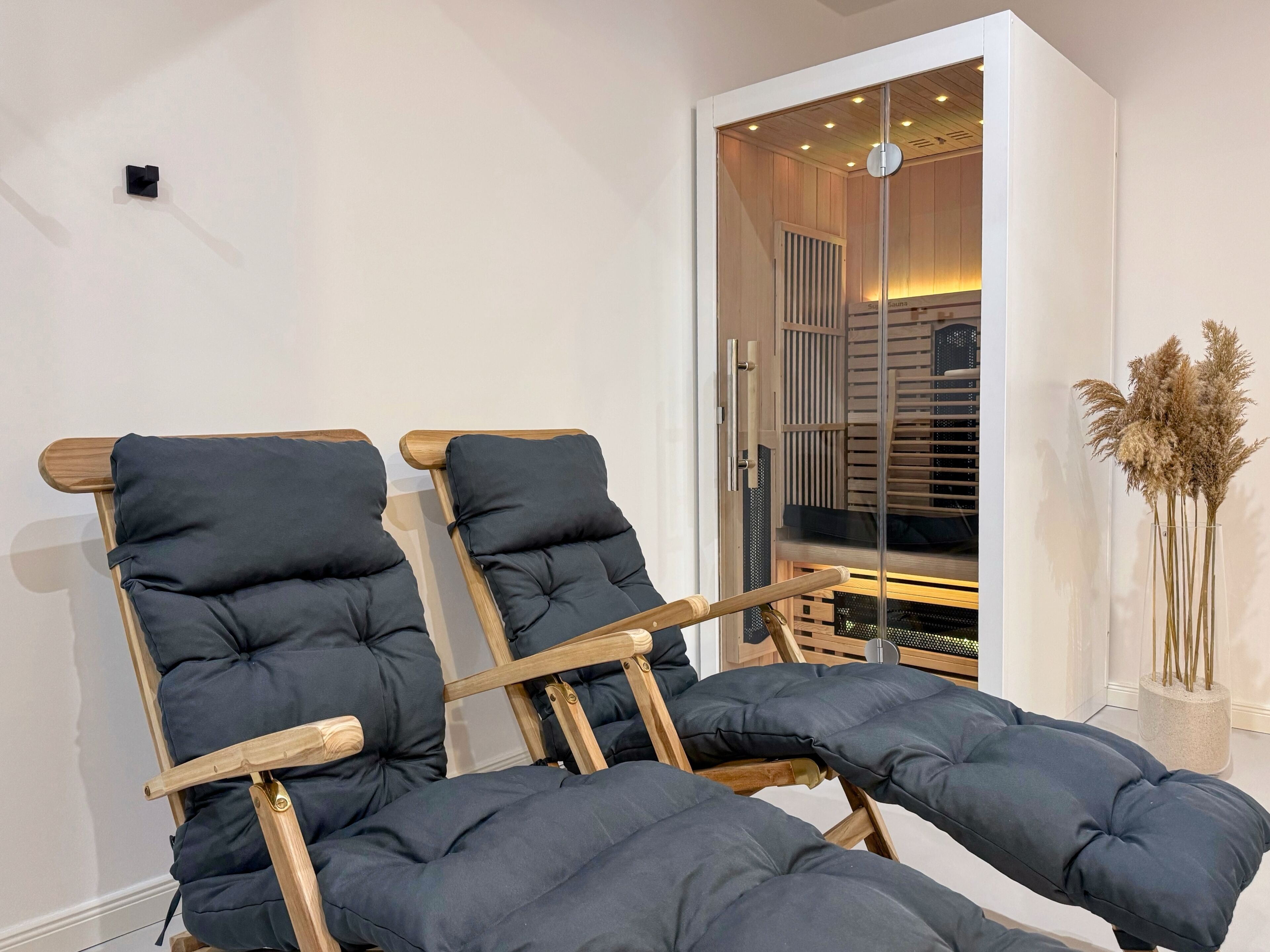 Una habitació amb dues cadires de fusta i seients foscos, una porta de vidre que dona a un interior de fusta, i un vas amb plantes seques sobre una taula.