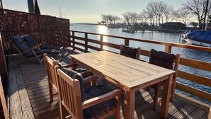 Een houten terras met een tafel en stoelen, uitkijkend op een waterpartij met een steiger op de achtergrond.