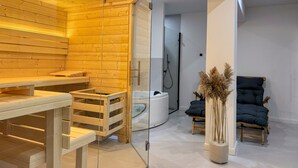 Een moderne spa-ruimte met houten lambrisering, een sauna met glazen wanden, een zwarte massagestoel en een klein tafeltje met een vaas gedroogde planten.