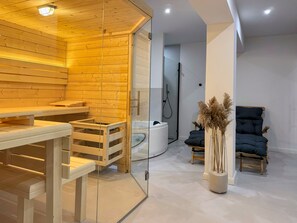Ein moderner Wellnessraum mit Holzvertäfelung, einer in Glas eingefassten Sauna, einem schwarzen Massagesessel und einem kleinen Tisch mit einer Vase getrockneter Pflanzen.