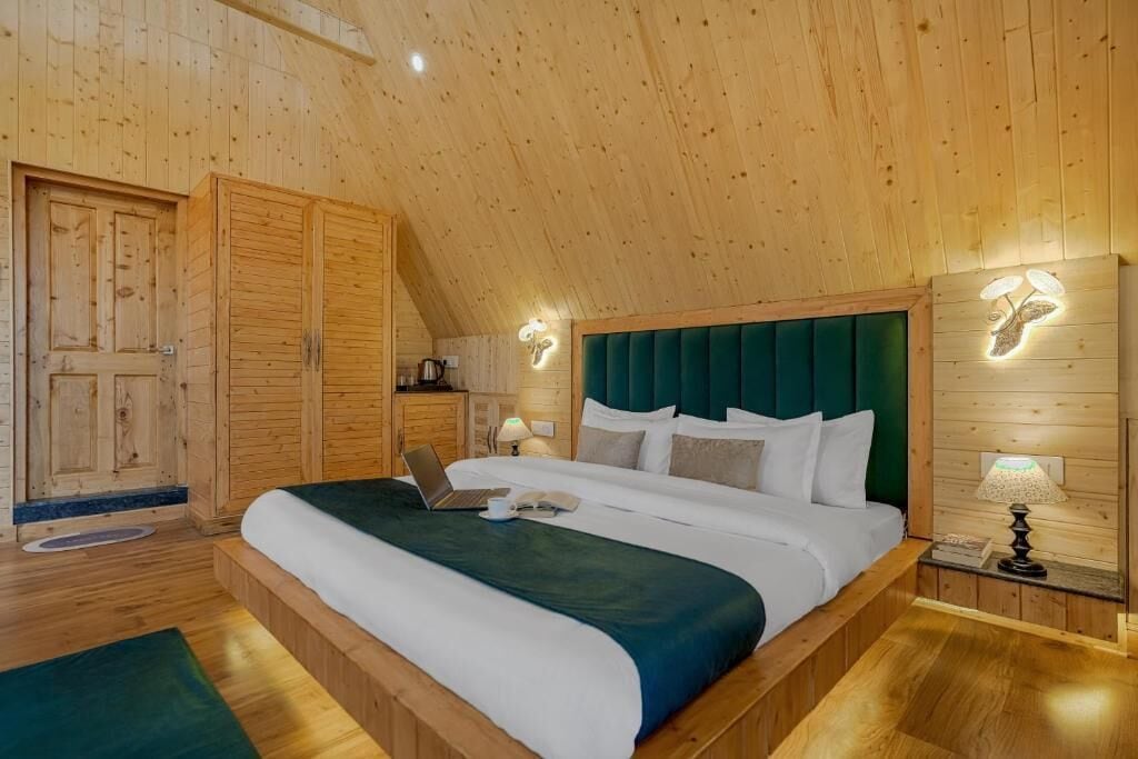 A bedroom with a wooden ceiling, a bed with a green headboard, a desk with a laptop, and a bedside lamp.