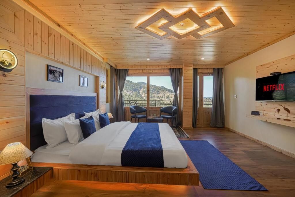 Habitación de hotel con cama, televisor, balcón con sillas y vistas a las montañas.