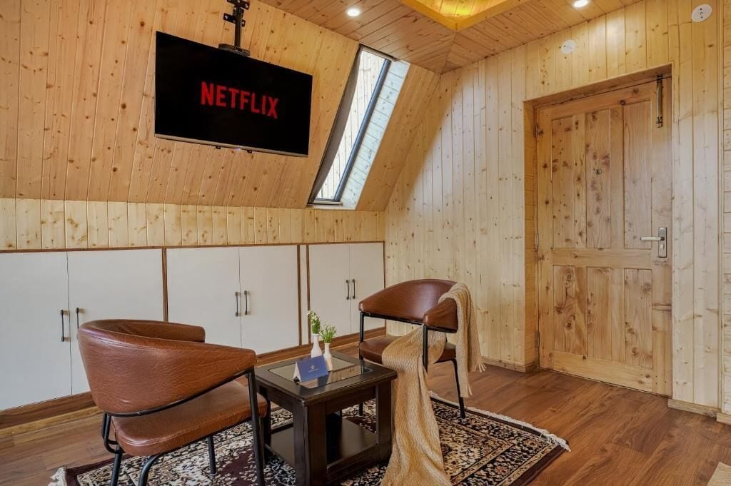 Ein Zimmer mit Holzwänden, einem an der Wand montierten Fernseher, zwei Ledersesseln, einem kleinen Tisch und einer Tür.