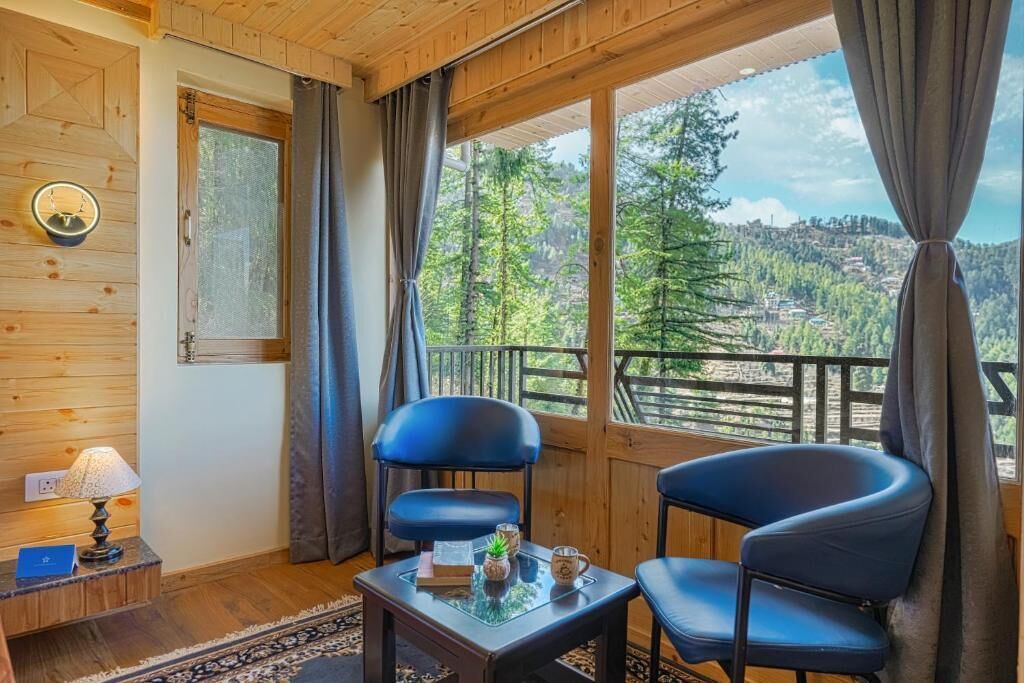 A room with wooden walls, a balcony with a view of a forested area, two black chairs, a small table with a lamp, and a window with curtains.