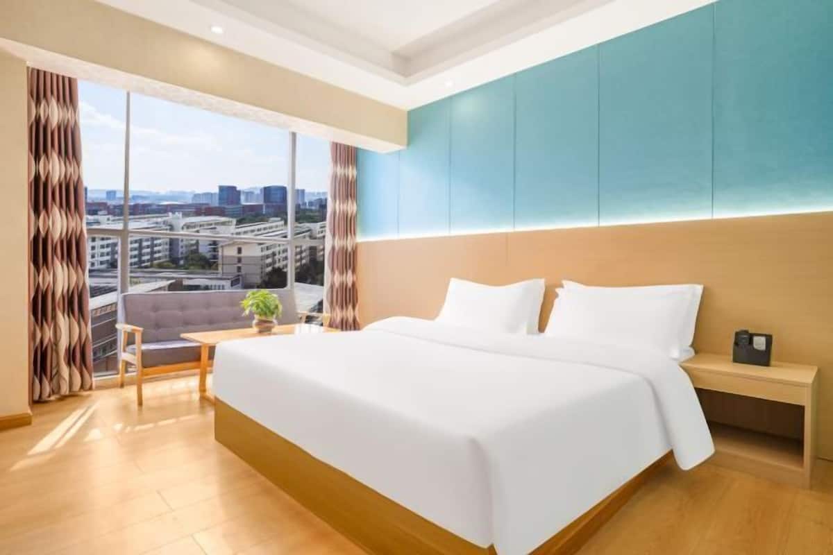 Habitación de hotel moderna con una cama grande, un escritorio pequeño, vistas a la ciudad y un ventanal amplio.
