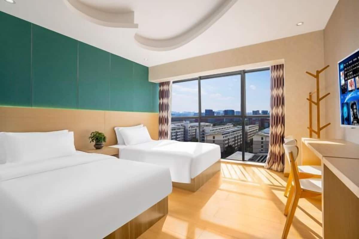 Habitación de hotel con dos camas, un ventanal grande con vista a la ciudad, una mesita con una planta y un televisor en la pared.