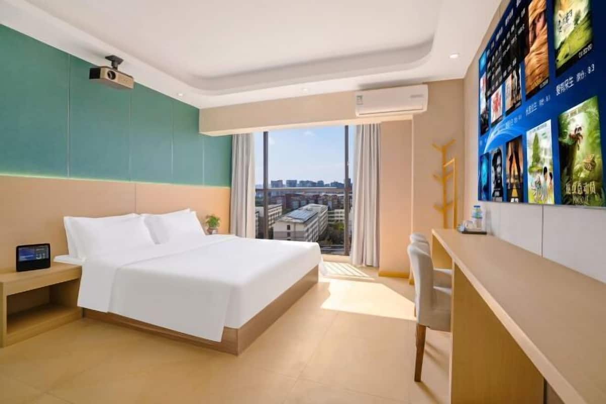 Una habitación de hotel con cama, mesita de noche, un escritorio con silla, un ventanal con cortinas y una pared con carteles de películas.