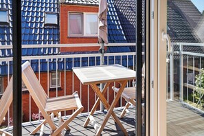 Ein Balkon mit Tisch und zwei Holzstühlen, eine rote Backsteinmauer und ein blau-gefliestes Dach.