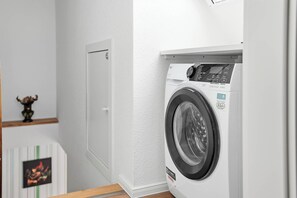 A compact, modern washing machine in a clean, white-walled laundry room with a door and a small decorative item on the wall. - New Platzhirsch vacation apartment in Greetsiel for a relaxing vacation (Greetsiel)