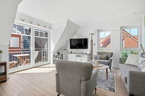 A spacious loft apartment with a large sliding glass door leading to a balcony, a flat-screen TV mounted on the wall, and a cozy seating area with a round coffee table. - New Platzhirsch vacation apartment in Greetsiel for a relaxing vacation (Greetsiel)