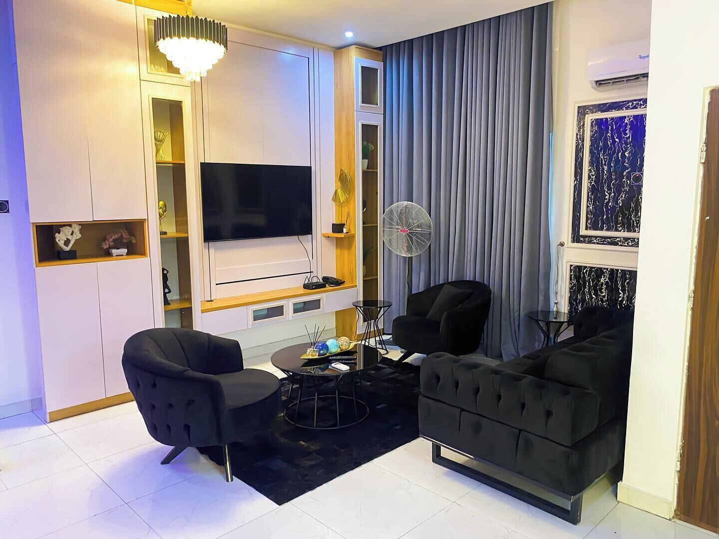 A modern living room with a flat-screen TV, two black sofas, a coffee table, a fan, and a decorative wall.