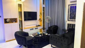 A modern living room with a flat-screen TV, two black sofas, a coffee table, a fan, and a decorative wall.