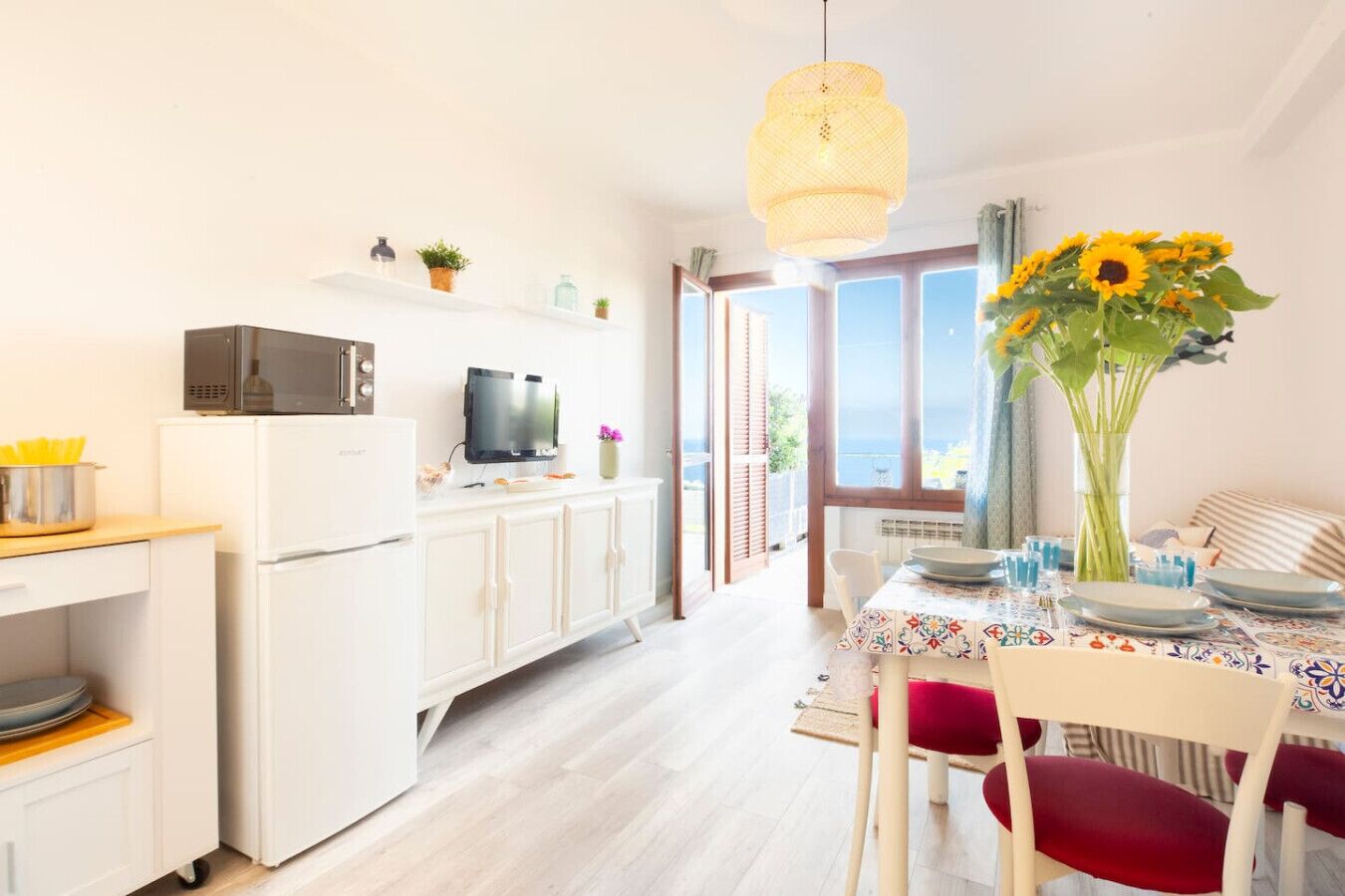 A modern kitchen with white cabinetry, a microwave, and a television. There is a dining area with a table set for two, a vase with sunflowers, and a view of the sea through large windows.