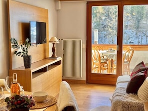 Una habitación acogedora con televisión, una mesa con una botella y vasos, una planta y vistas a un balcón con sillas.