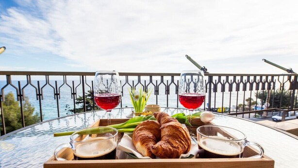 Bandeja de café da manhã com croissants, dois copos de vinho tinto e um copo de vinho branco numa varanda com vista para o mar.