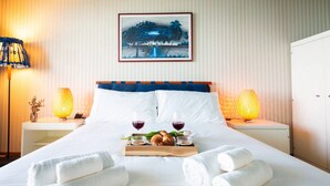 Quarto de hotel com uma cama, duas luminárias laterais, um tabuleiro com pão e copos de vinho, e um quadro na parede.
