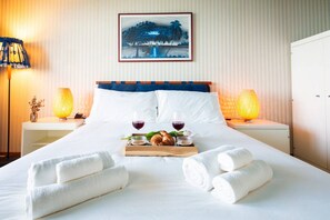 Ein Hotelzimmer mit einem Bett, zwei Nachttischlampen, einem Tablett mit Brot und Weingläsern sowie einem Bild an der Wand.