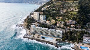 Vista aérea de um hotel à beira-mar, com vários edifícios, piscina e costa rochosa.