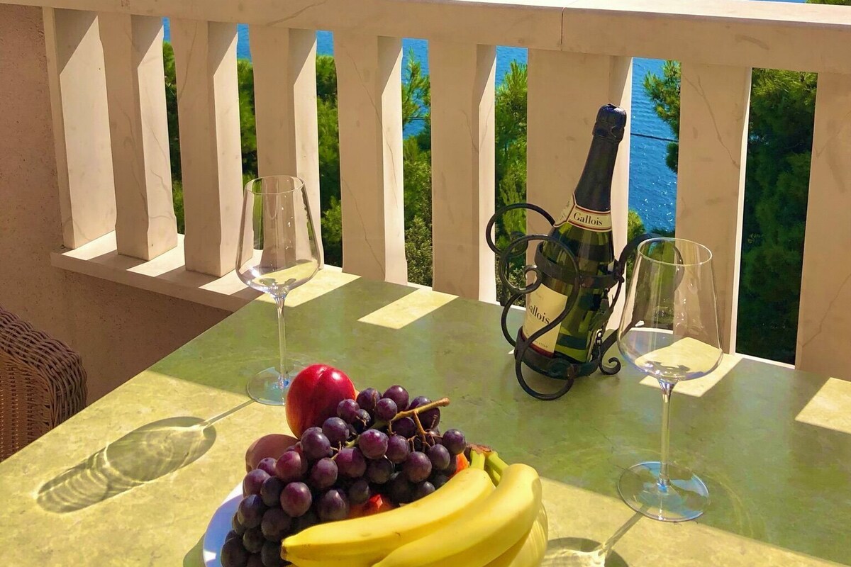 A table with a bottle of wine, glasses, a plate of fruit, and a scenic view of a body of water and mountains.