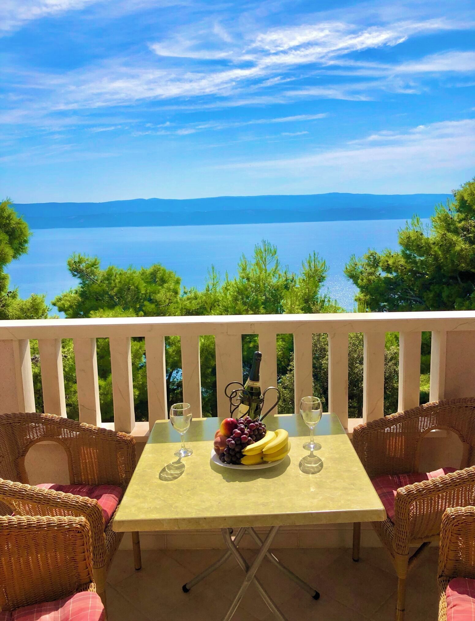 A table with a bottle of champagne, a fruit platter, and two glasses on a balcony overlooking a body of water and distant mountains.