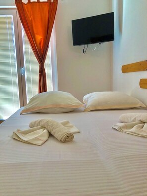 A neatly made bed with pillows and rolled towels, a television mounted on the wall, and a window with blinds. - Apartments Luka (Omis)