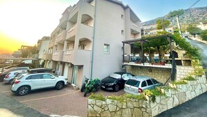 A multi-story building with balconies, a parking area with cars and a motorcycle, and a stone wall with greenery. - Apartments Luka (Omis)